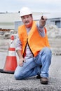 Man wearing hard hat and safety vest kneeling next to traffic cone and pointing Royalty Free Stock Photo