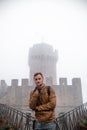 Man wearing leather jacket and backpack. Fortification, old castle on background. Journey. Mystical atmosphere, fog, white haze,