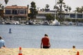 A man wearing a red shirt standing in the sand at Horny Corner Beach with luxury homes and boats and yachts docked