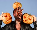 Man wearing scary makeup holds pumpkin on head on blue background. Demon with horns and scary face licks carved jack