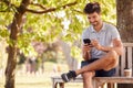 Man Wearing Summer Shorts Sitting On Park Bench Under Tree With Takeaway Coffee Using Mobile Phone