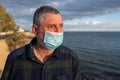 A man is wearing a surgical mask and looks sad and frustrated at the sea.