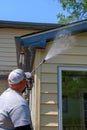 Man wearing a white baseball cap power washing the siding and roof of a beige and blue house.