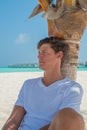 Man wearing white t-shirt and black shorts relaxing under palm tree at tropical sandy beach at island luxury resort Royalty Free Stock Photo