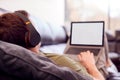 Man Wearing Wireless Headphones Listening To Music Steaming From Laptop On Sofa At Home In Pyjamas Royalty Free Stock Photo