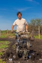 Man in wellingtons with cultivator ploughing ground in sunny day. Farmer plowing kitchen-garden in suburb. Land cultivation, soil