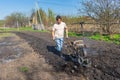 Man in wellingtons with cultivator ploughs ground in sunny day. Land cultivation, soil tillage. Spring work in garden. Gardening