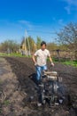 Man in wellingtons with cultivator ploughs ground in sunny day. Land cultivation, soil tillage. Spring work in garden. Gardening