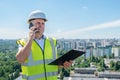 Man in a white helmet at a construction site holds a clipboard in his hands