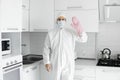 Man in white protective suit and in face medical mask and in a pink oven gloves cooks in the white kitchen at home