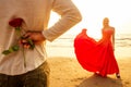 Man in a white shirt giving roses to surprised wonder woman in a long red dress on the ocean beach. romantic date Royalty Free Stock Photo