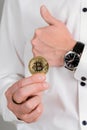 A man in a white shirt holds a bitcoin gold coin in his hand and shows a thumbs up gesture Royalty Free Stock Photo