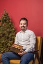 Man in white shirt holds Christmas gift gold box on red background and Christmas tree Royalty Free Stock Photo