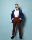 Man in white shirt, jacket, brown pants, black loafers. Straightening sunglasses, hand in pocket, posing on blue background