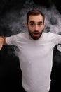 Man in white t-shirt on black background releasing a cloud of smoke
