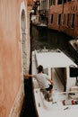 A man in a white T-shirt stands in a boat and rests his hand on the wall. Gondoliers sail boats.