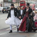A man in a white wedding dress and a top hat with a woman in a gothic red velvet dress walking down the street in London Bridge