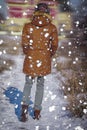 A man in a winter hat and fur coat walks away along the path with his back turned, it is snowing outside