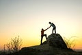 Man and woman hikers helping each other to climb a big stone at sunset in mountains. Couple climbing on a high rock in evening