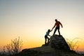 Man and woman hikers helping each other to climb a big stone at sunset in mountains. Couple climbing on a high rock in evening