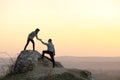 Man and woman hikers helping each other to climb stone at sunset in mountains. Couple climbing on high rock in evening nature.