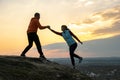 Man and woman hikers helping each other to climb stone at sunset in mountains. Couple climbing on high rock in evening nature.