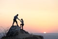 Man and woman hikers helping each other to climb stone at sunset in mountains. Couple climbing on high rock in evening nature.