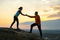 Man and woman hikers helping each other to climb stone at sunset in mountains. Couple climbing on high rock in evening nature.