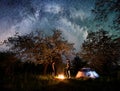 Man and woman hikers standing at a campfire near tent under trees and night sky full of stars and milky way Royalty Free Stock Photo