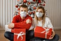 Man and woman in protective masks on faces are sitting near Christmas tree at home holding present gifts and looking at camera. Royalty Free Stock Photo