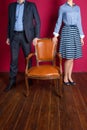 Man and woman standing around empty armchair.