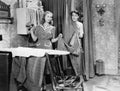 Man and woman standing in a kitchen while she is ironing his pants and he is behind a curtain