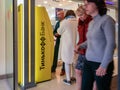 A man and a woman are standing near the yellow terminal with the Tinkoff Bank logo. ATM at the entrance to the mall. Visitors pass