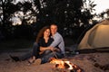 Man and woman tourists sitting in front of tent at campfire and forest background. Night camping on shore. Active