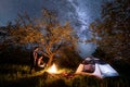 Man and woman tourists standing at a campfire near tent under trees and beautiful night sky full of stars and milky way Royalty Free Stock Photo