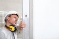 Man worker looks at the spirit level checks the wall with hard hat and ear protection headphones, white wall with copy space