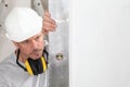 Man worker looks at the spirit level checks the wall with hard hat and ear protection headphones, white wall with copy space