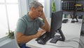 Man working in office looks stressed while using computer with one hand on his face, sitting at a modern clean workstation Royalty Free Stock Photo