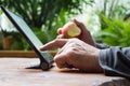 Man working on tablet computer with bitten apple in his hand. Wooden table. Green garden background. Royalty Free Stock Photo