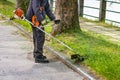 Man with worn bush cutter trims overgrown lawn round trees next to road