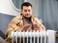 Man wrapped in cover warming hands at radiator in apartment