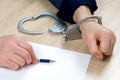 A man writes a confession at the police station. One hand is cuffed, the other is unbuttoned, pen in hand for writing explanations