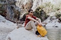 Man with yellow backpack sits on a riverside rock in the canyon with mountain cliffs in the background. Adult man taking