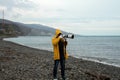 A man in a yellow raincoat takes photos from the seashore with a long-focus lens. It uses a monopod tripod to support the camera a