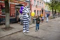 A man in a zebra costume on a city street greets passers-by and gives balloons to children. Advertising on the street