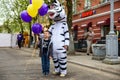 A man in a zebra costume on a city street greets passers-by and gives balloons to children. Advertising on the street