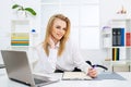 Managing Business Work. Happy young business woman using pc at office. Gorgeous blond secretary woman writing down notes Royalty Free Stock Photo