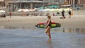 Woman tourist entering the sea with a surfboard on the beach in front of the village during the day Royalty Free Stock Photo