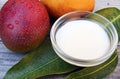 Mango body butter in a glass bowl and fresh ripe organic mango fruit and leaves on old wooden background.