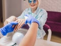 Manicure master in blue gloves creaming hands of elderly stylish woman in blue sunglasses and jeans jacket sitting at manicure sal